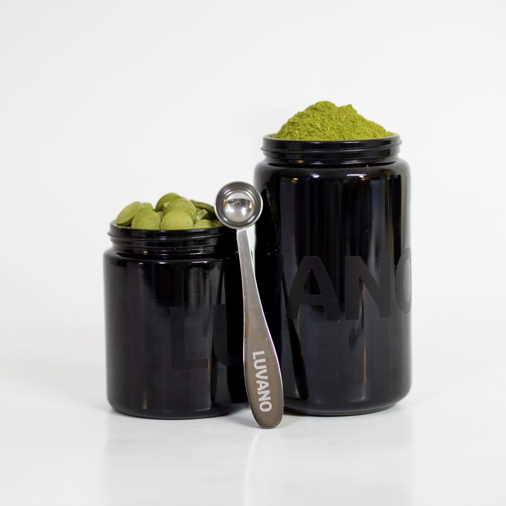 Zwei schwarze Luvano Violettglasbehälter, einer mit grünem Pulver, einer mit grünen Tabletten, daneben ein Dosierlöffel