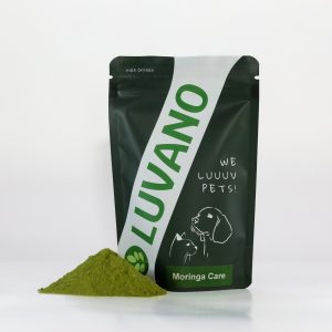 Grüner Luvano Pouchbag mit Ergänzungsfuttermittel für Hunde „Moringa Care“, davor grünes Pulver auf weißem Untergrund
